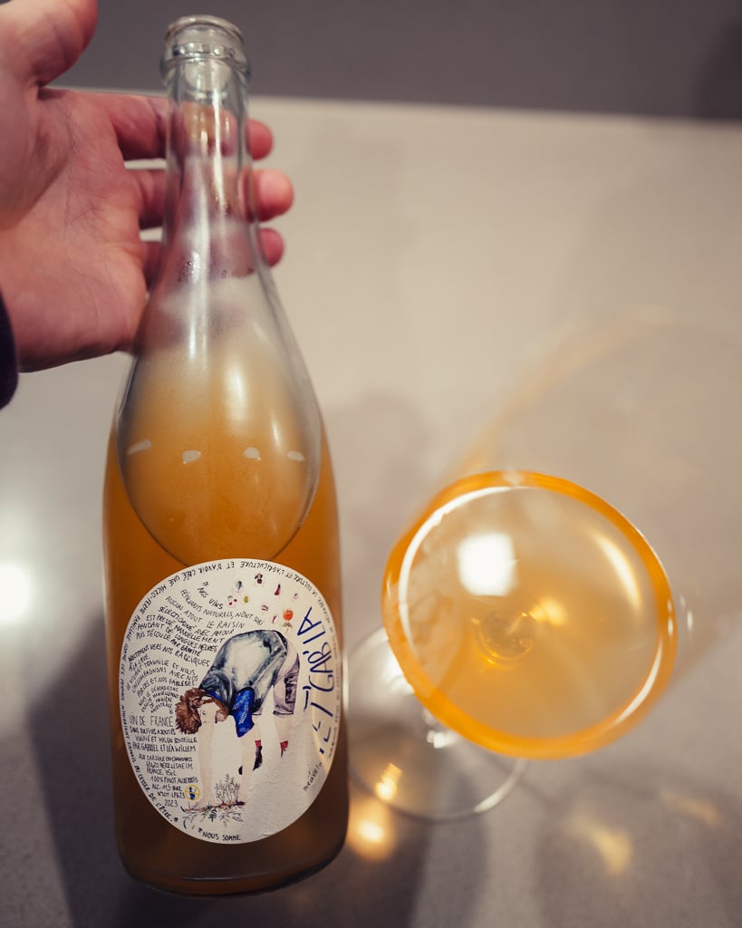 Hand holding the cloudy Pet'Gab'La pet-nat bottle beside a glass of hazy orange wine