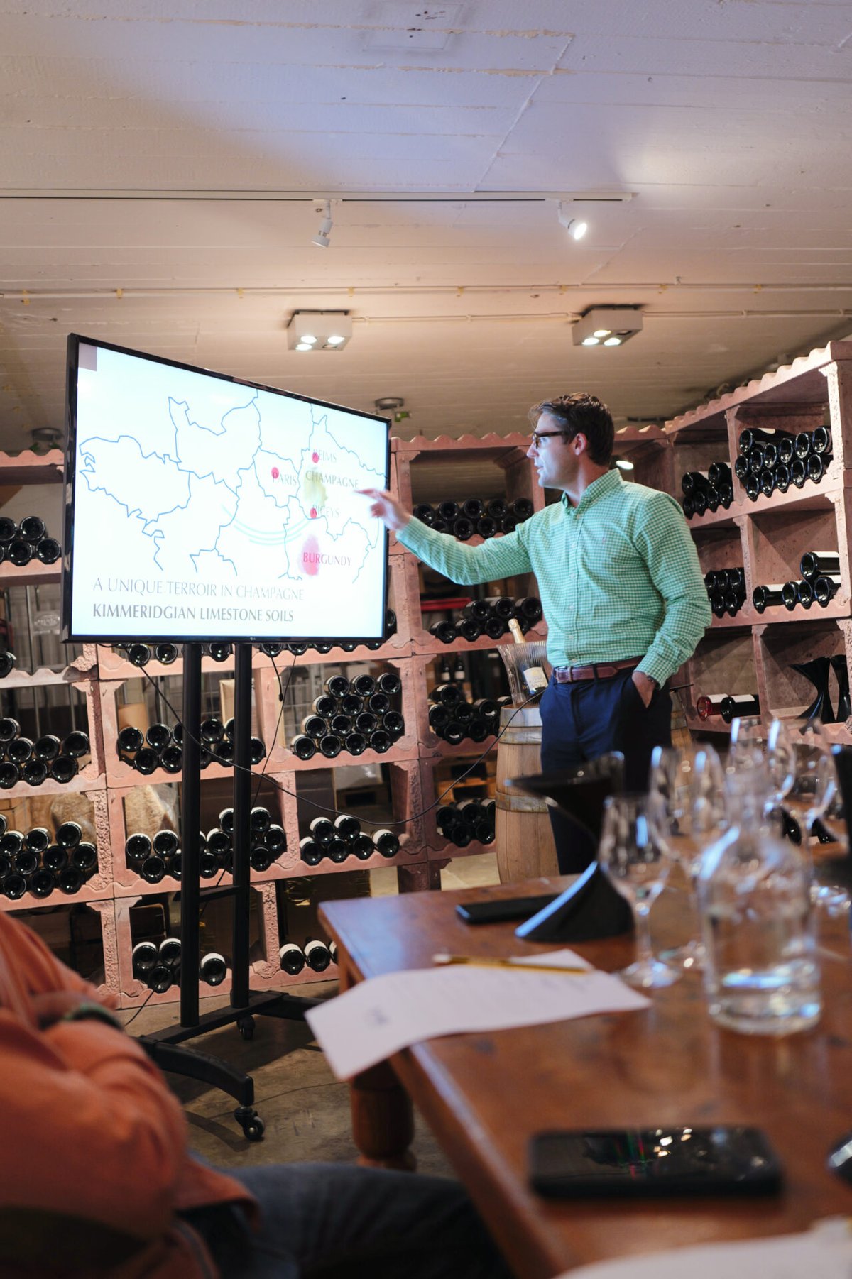 Speaker pointing at a map on a screen during a masterclass in a wine cellar with bottle racks and tasting glasses