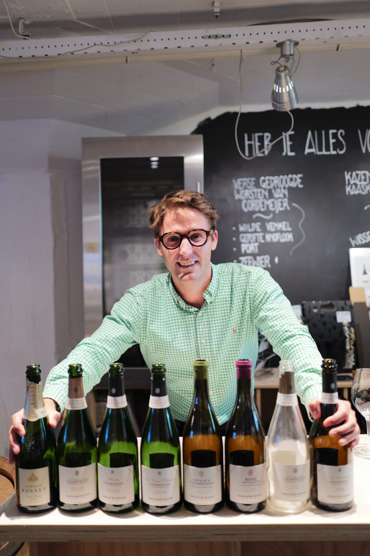 Man in a green checked shirt standing behind a row of eight Champagne and wine bottles in a wine shop