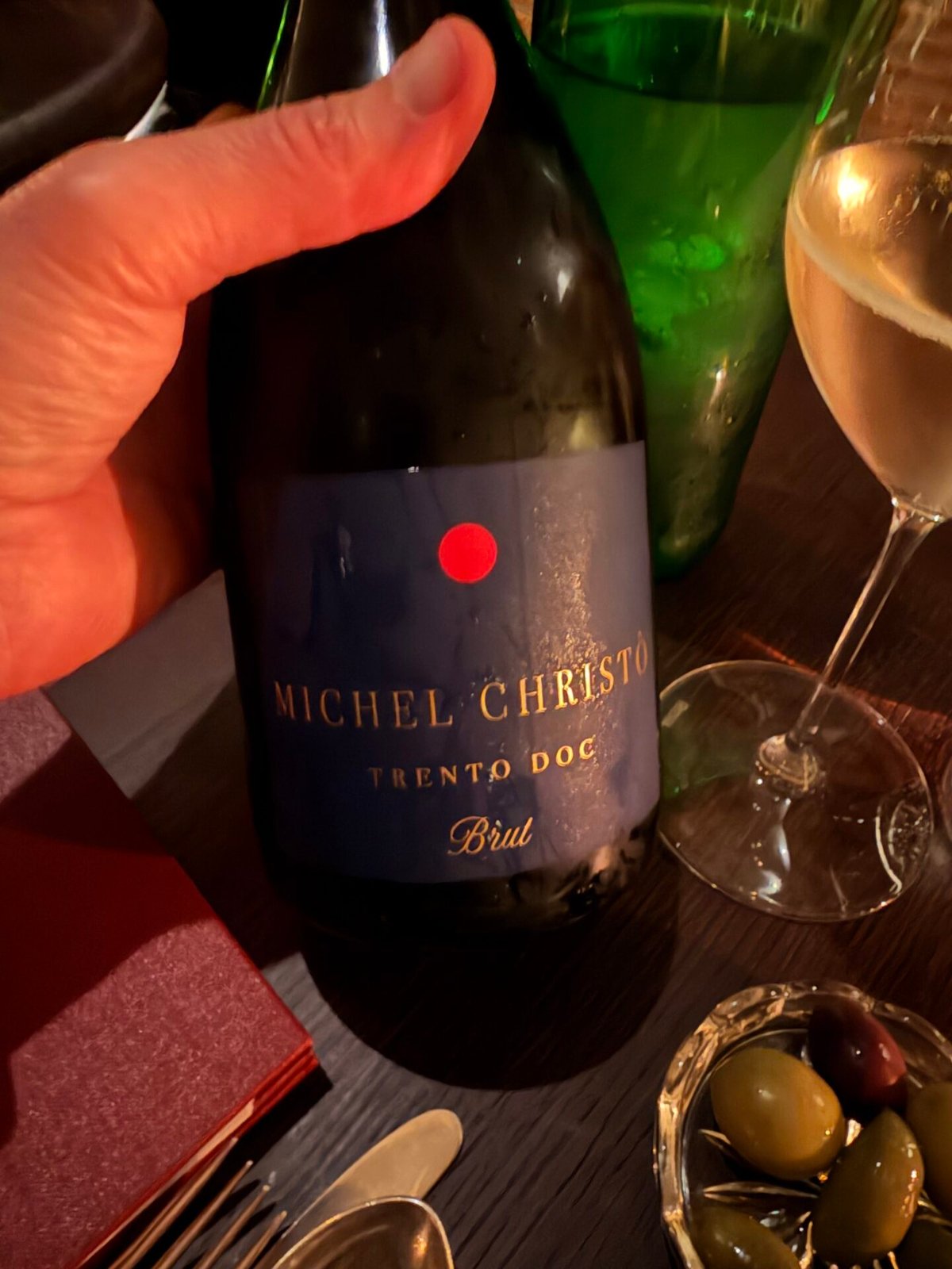 Hand holding a Michel Christò Trento DOC sparkling bottle at a dim restaurant table with olives