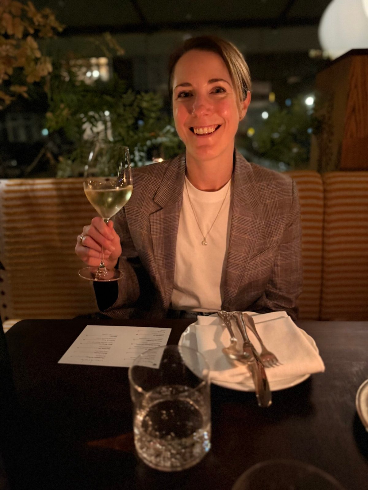 Woman in a grey blazer smiling and holding a glass of white wine at a restaurant table