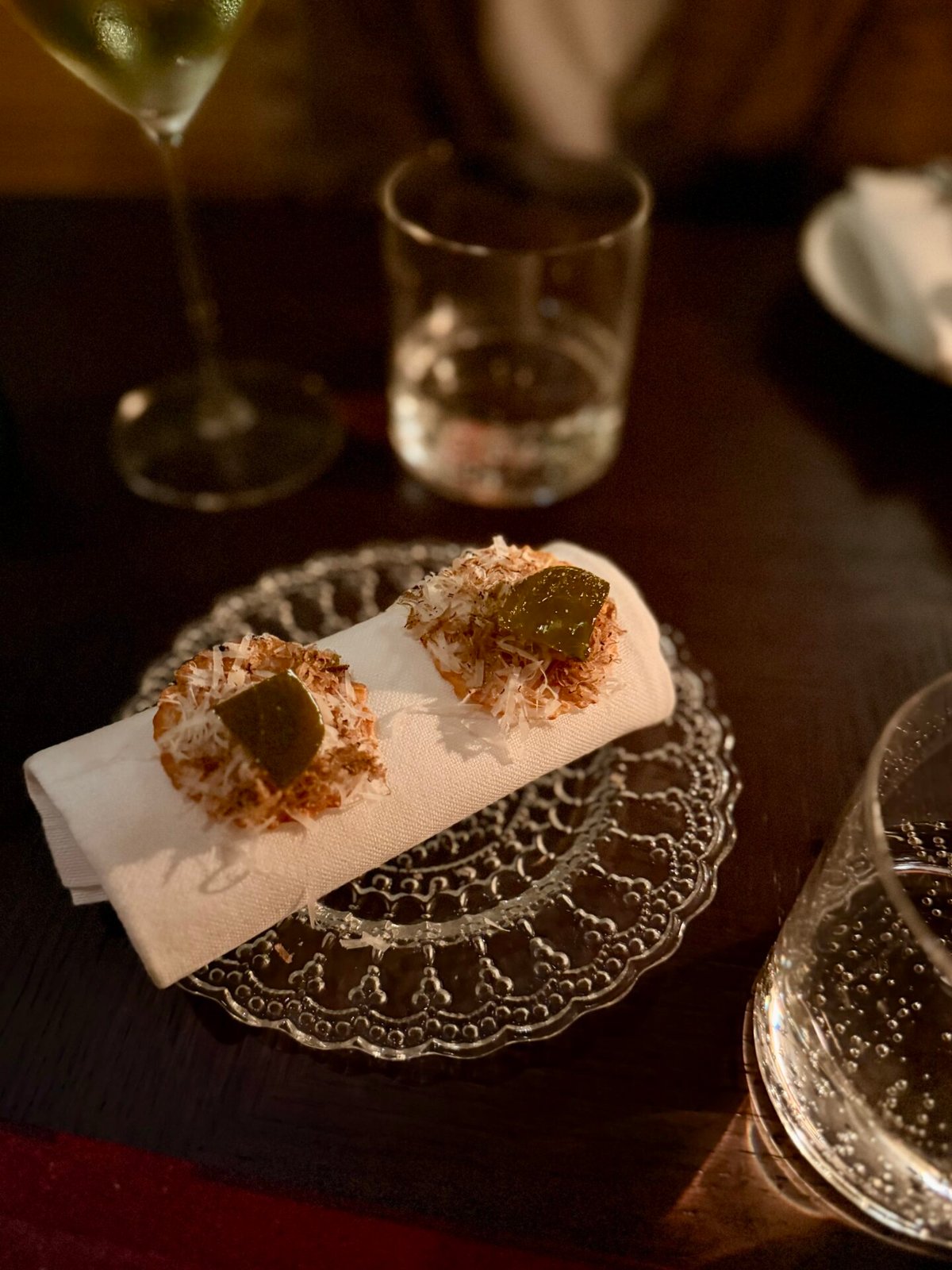 Two small amuse-bouche bites on a lace glass plate beside a sparkling wine glass