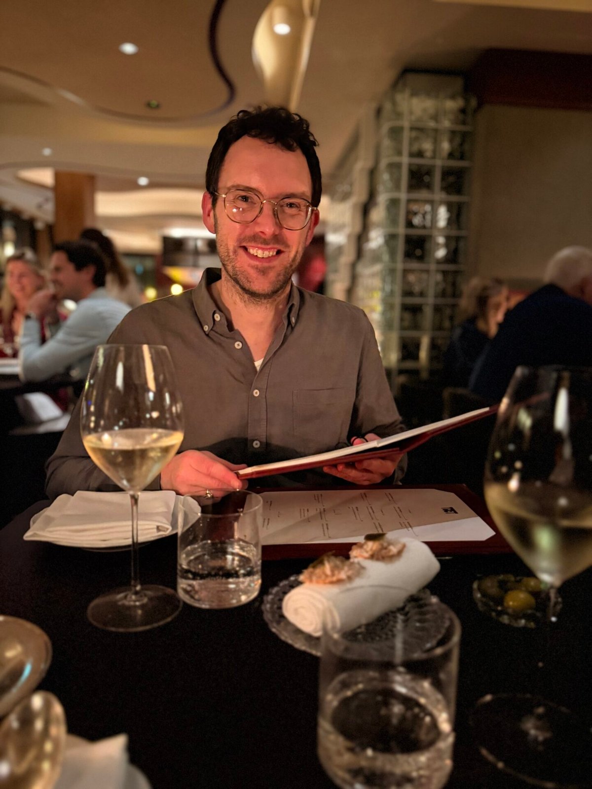 Smiling man holding a menu at a restaurant table with white wine and an amuse plate