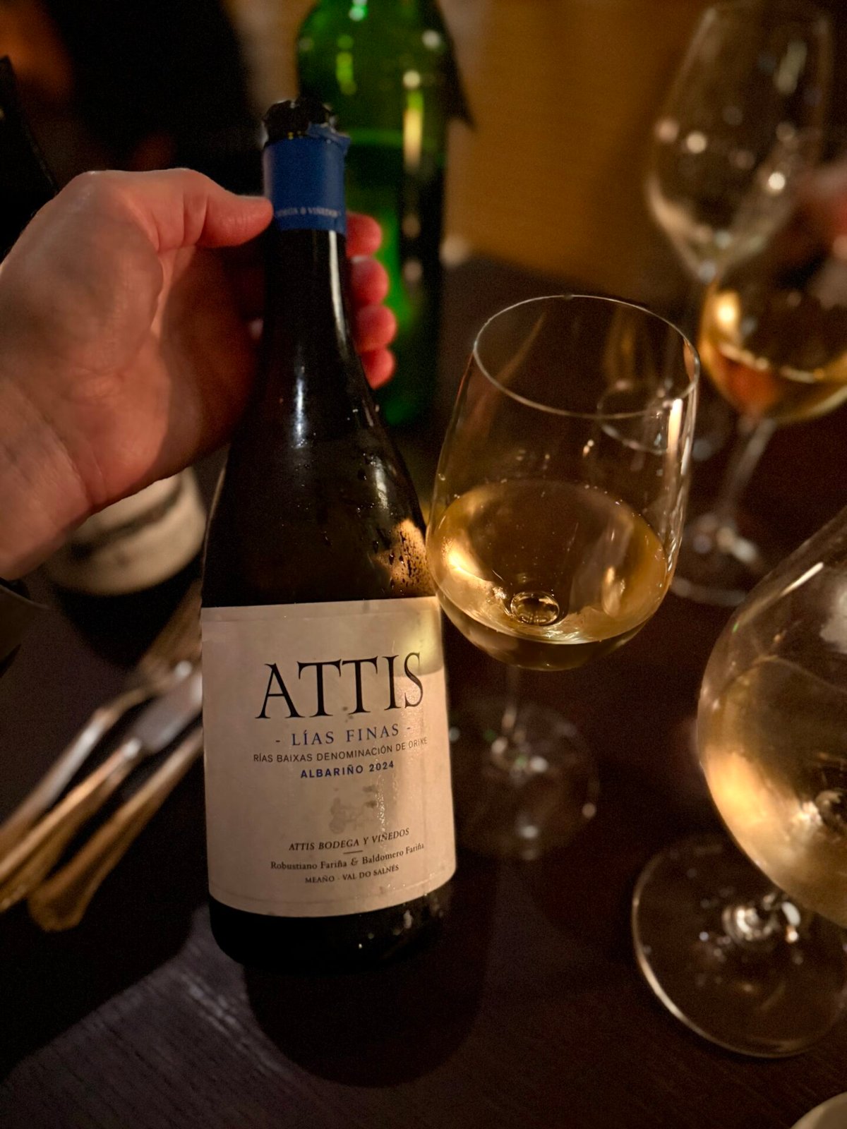 Hand holding an Attis Lías Finas Albariño bottle beside a glass of white wine in dim light