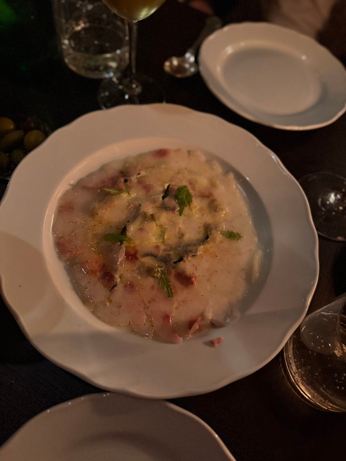 Creamy dish with herbs in a white bowl on a dark table beside a wine glass in low light