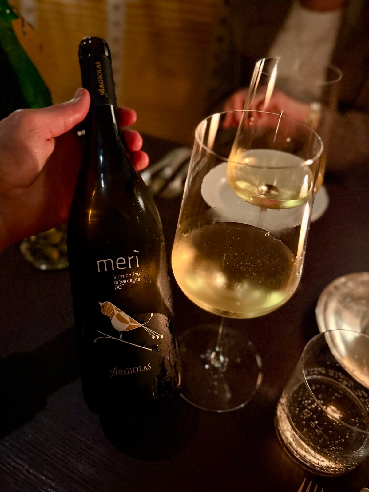 Hand holding a Meri Argiolas Vermentino bottle beside a glass of white wine at the table