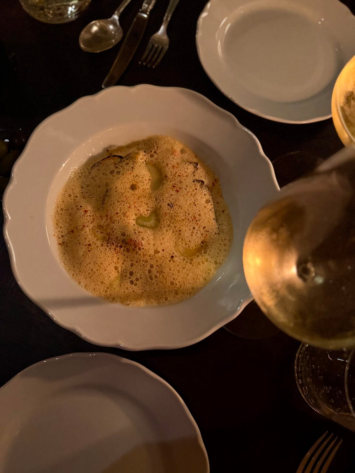 Foamy soup in a white bowl on a dark restaurant table beside a glass of white wine