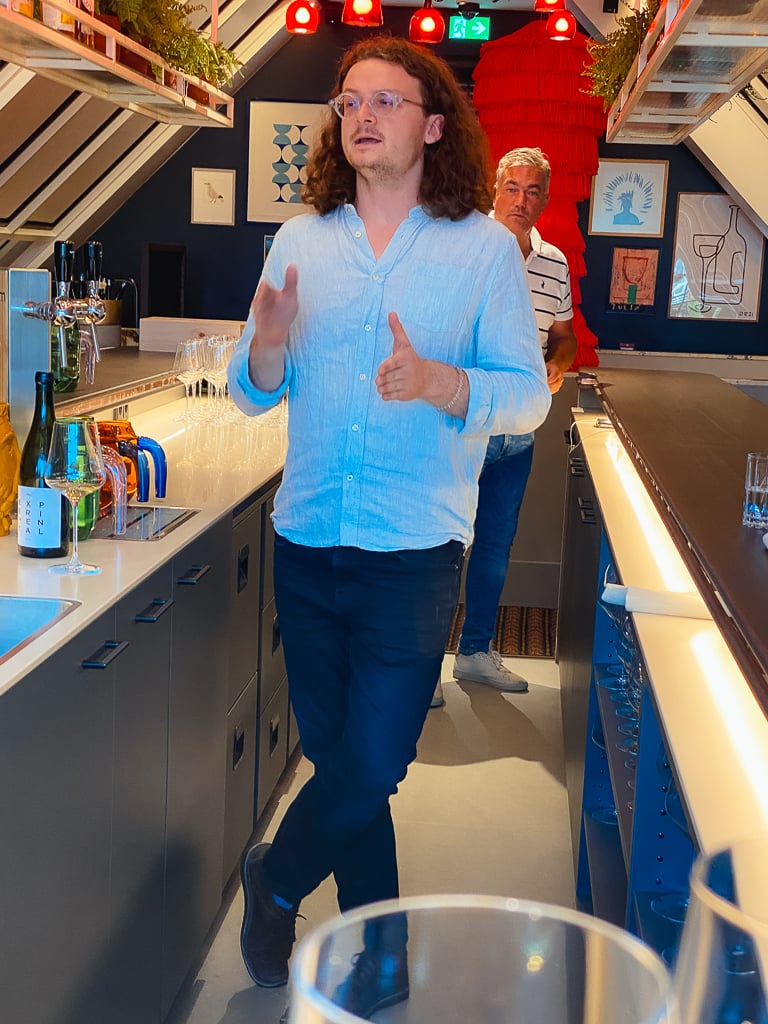 Winemaker Sam Lambson gesturing and talking while standing in a bar with bottles behind him