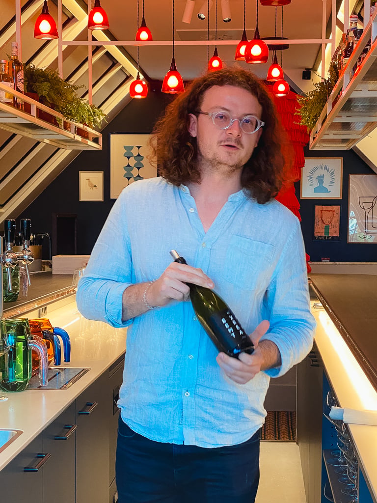 Winemaker Sam Lambson in a blue shirt holding a wine bottle in a bar under red pendant lights