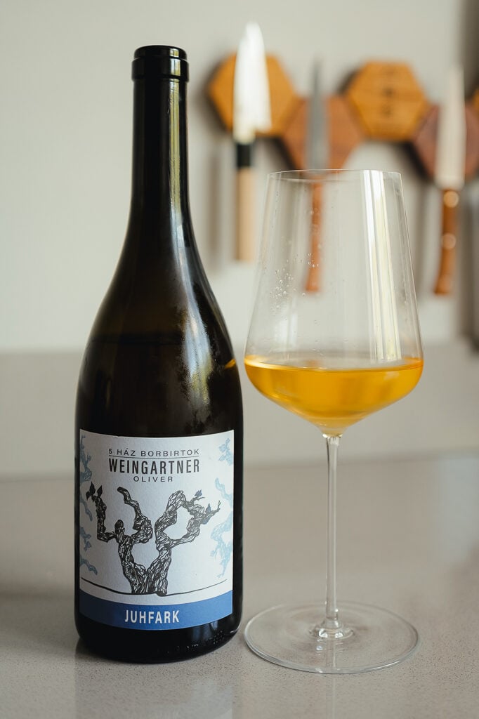Weingartner Juhfark bottle with blue label beside a glass of amber orange wine