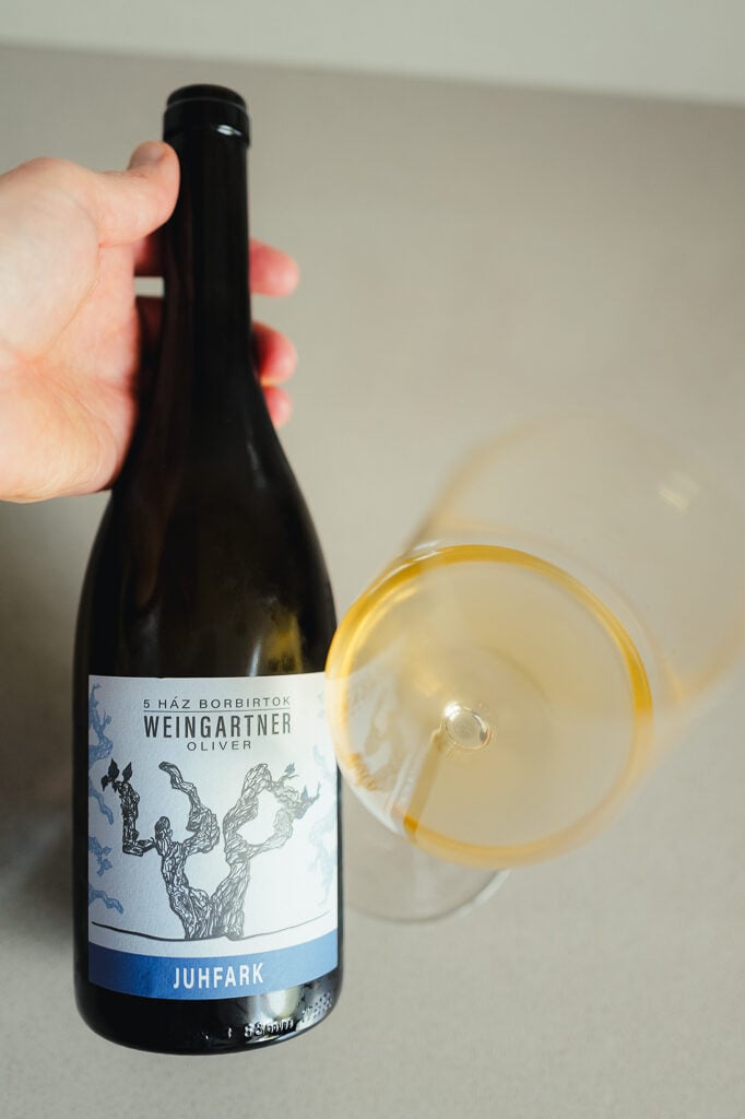 Hand holding a Weingartner Juhfark bottle next to a glass of amber orange wine