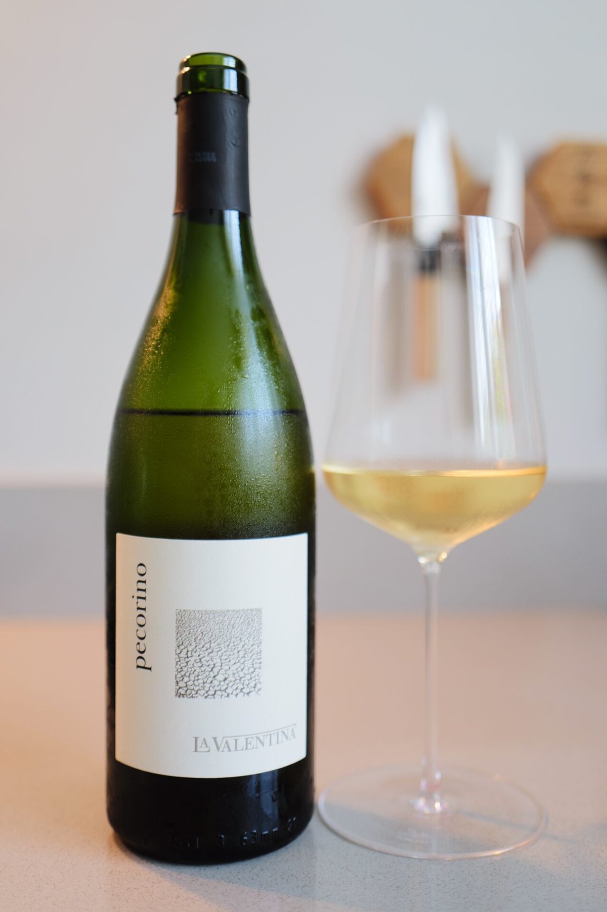 La Valentina Pecorino bottle next to a glass of golden white wine on a pale countertop