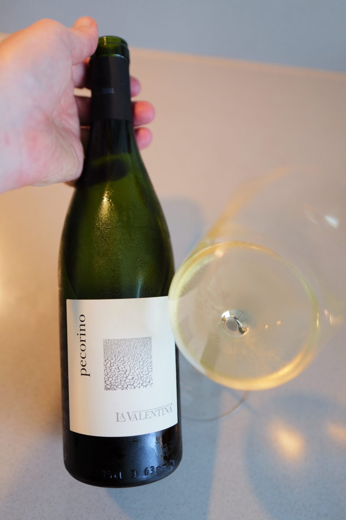 Hand holding a green La Valentina Pecorino bottle beside a glass of pale gold white wine