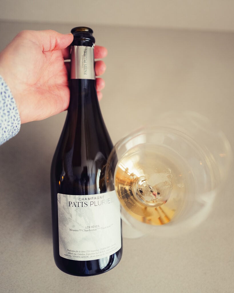 Hand holding a Patis Pluriel Les Sèves bottle next to a tilted glass of amber champagne