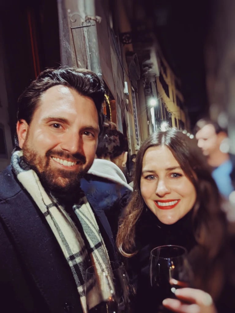 Smiling couple Lauren and Spencer holding wine glasses in a narrow lamplit street at night