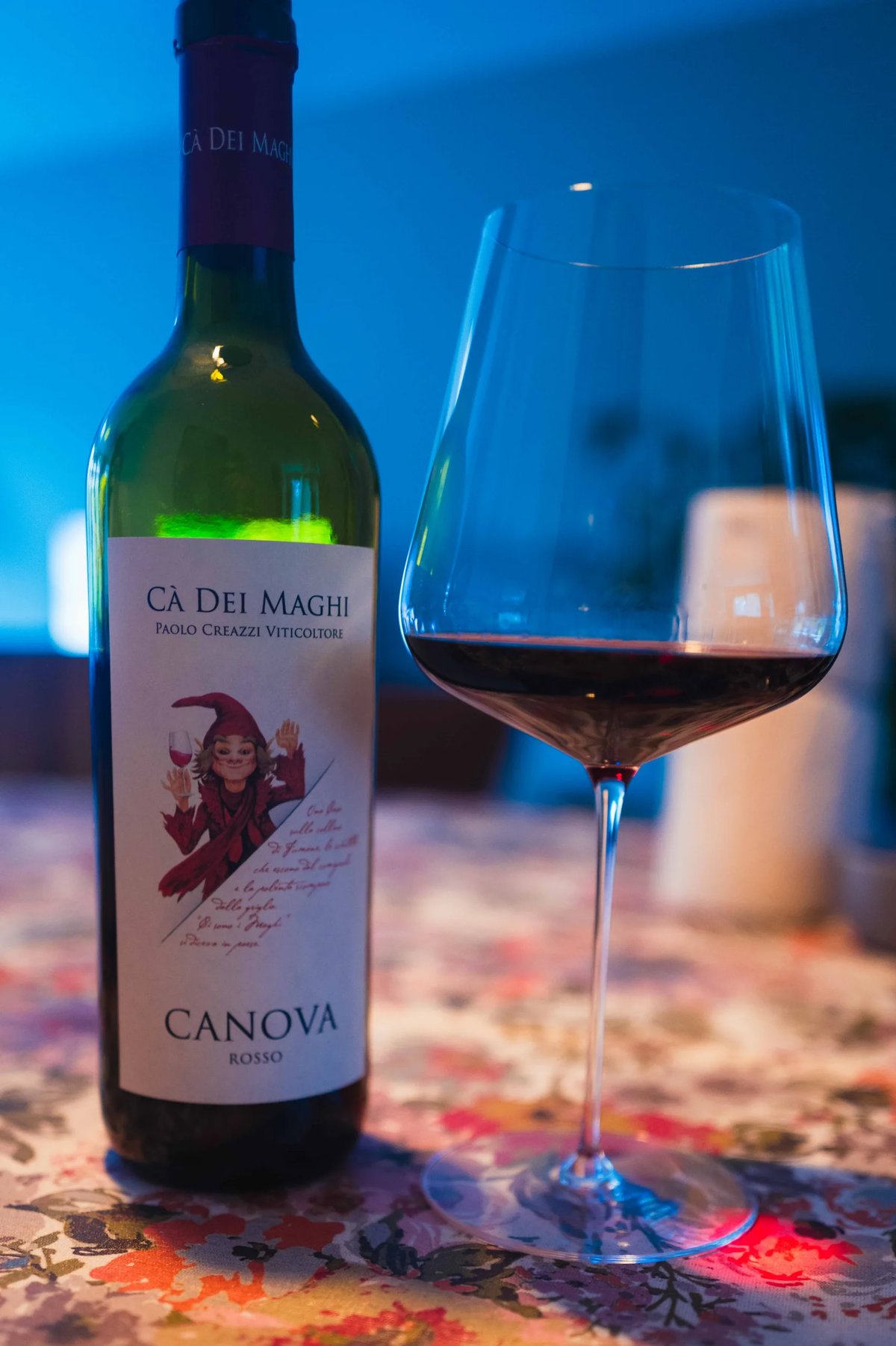 Cà Dei Maghi Canova Rosso bottle with a gnome label beside a glass of red wine in blue light