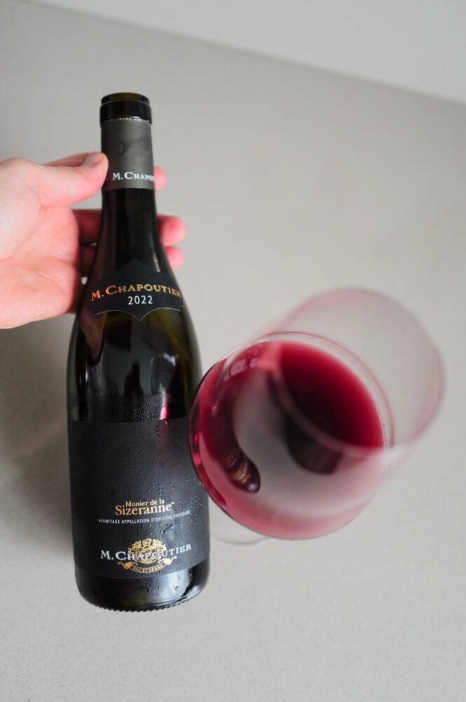 Hand holding the M. Chapoutier Sizeranne Hermitage bottle beside a tilted glass of red wine