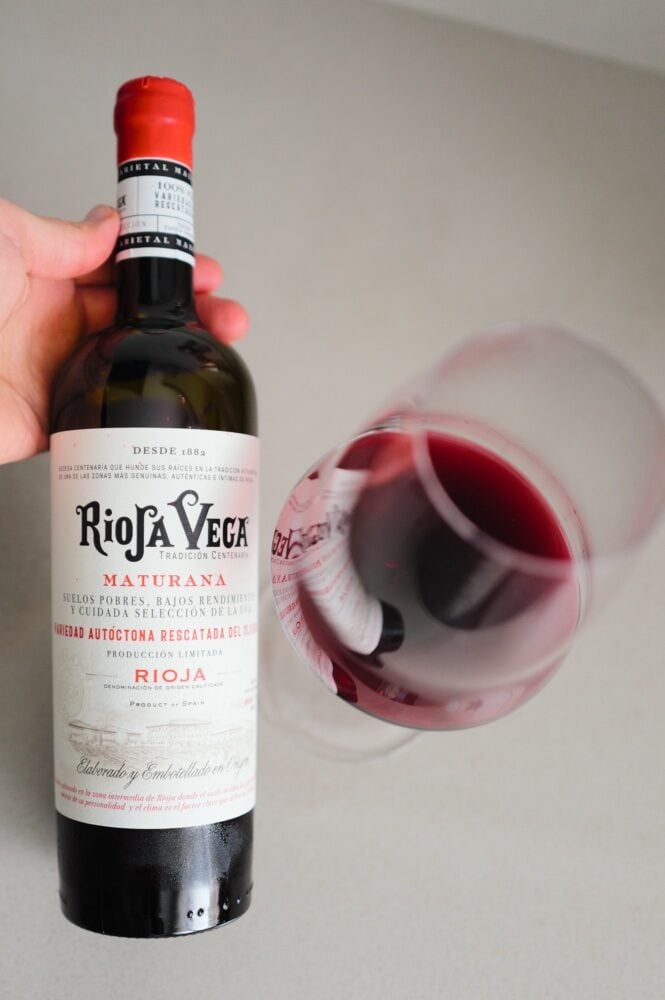 Hand holding the Rioja Vega Maturana bottle beside a tilted glass of dark red wine