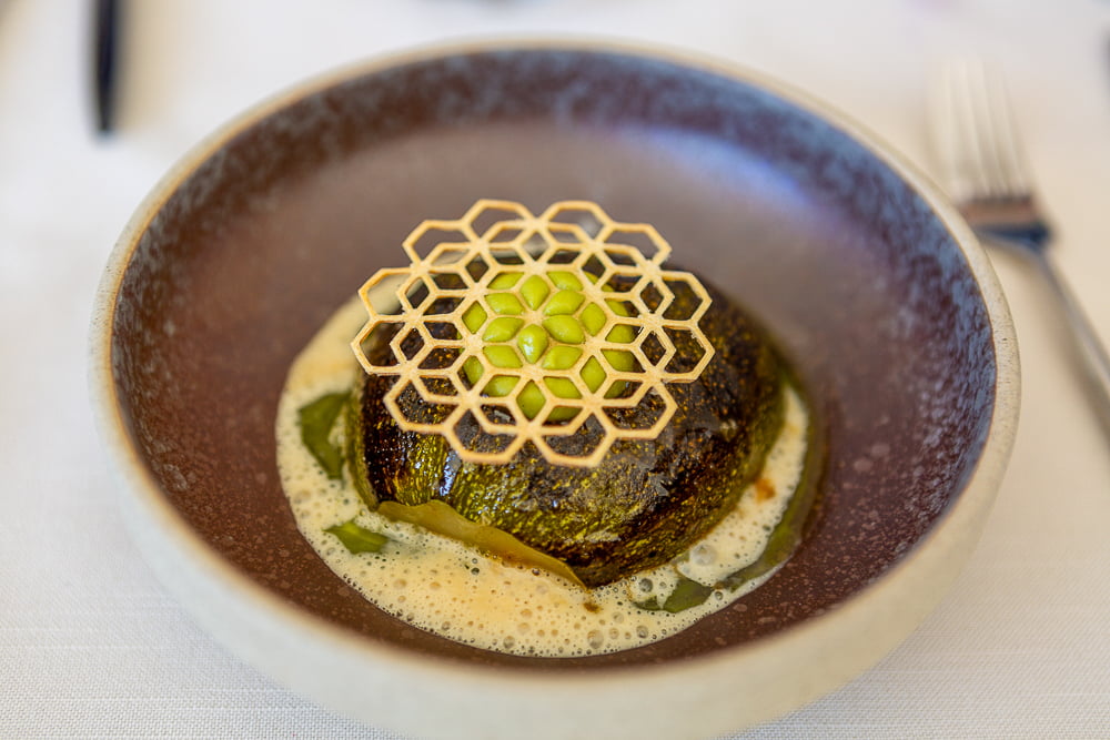 Grilled whole courgette topped with a honeycomb tuile in a frothy sauce in a brown bowl