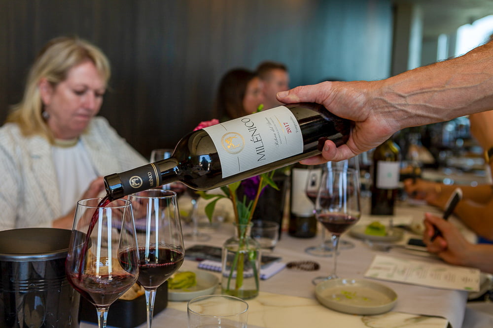 Hand pouring Milénico Ribera del Duero red into glasses at a tasting table with guests