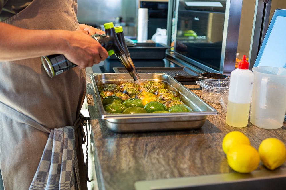 Chef using a blowtorch on a tray of halved courgettes in a kitchen with lemons nearby