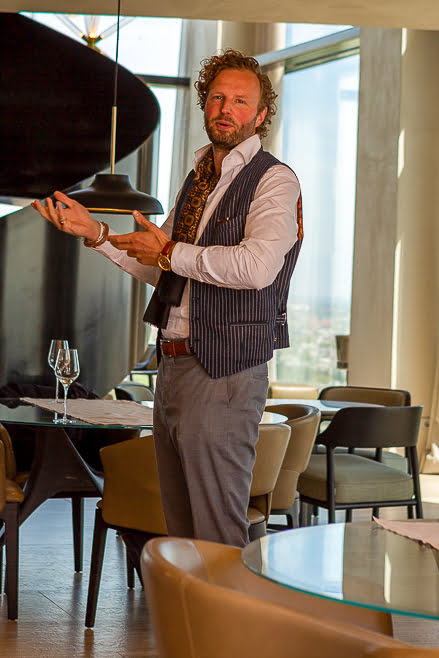 Man in a striped waistcoat and cravat presenting at a high-rise restaurant with city views