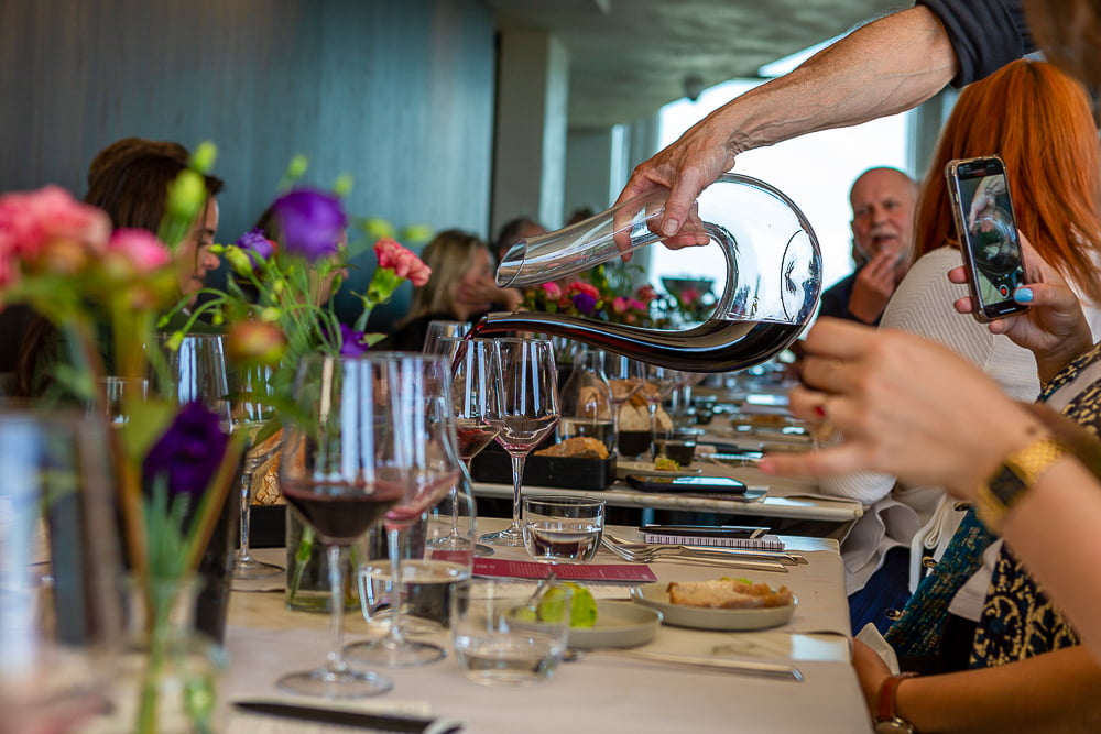 Hand pouring red wine from a silver decanter at a long tasting table while a guest films