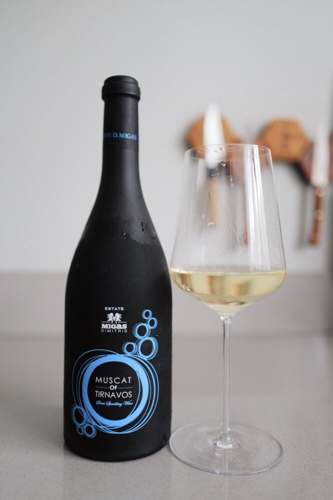 Black Migas Muscat of Tirnavos sparkling bottle next to a glass of golden wine
