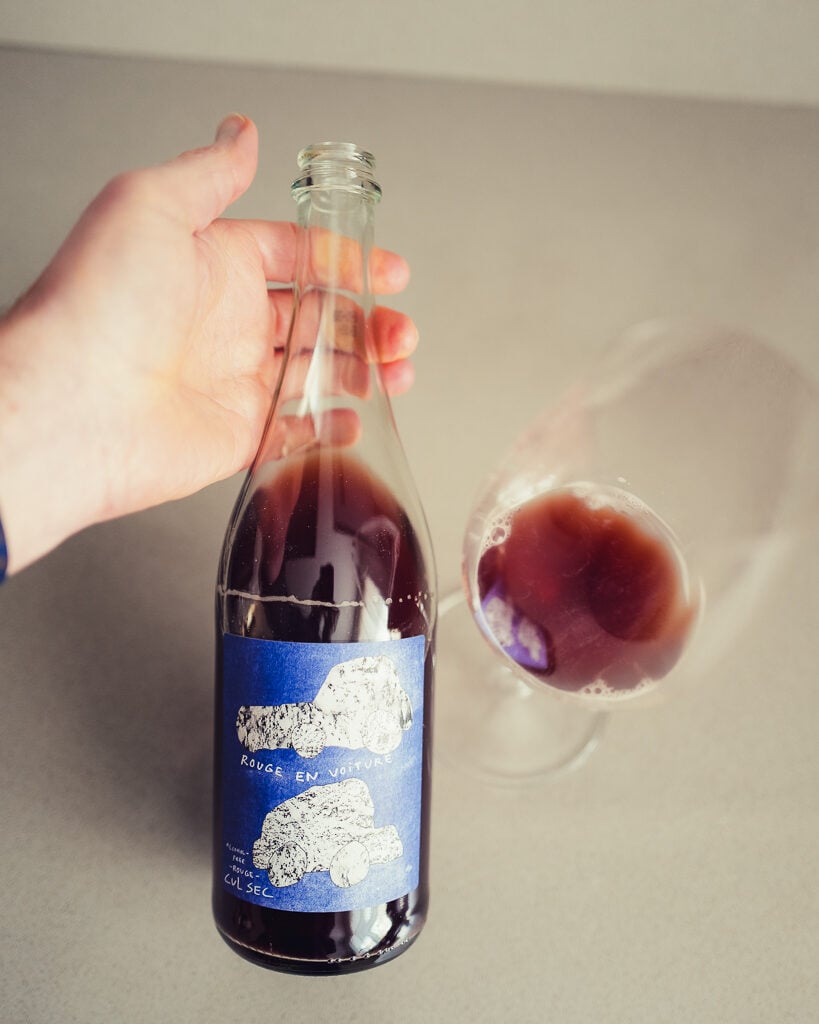 Hand holding the blue-labelled Rouge en Voiture Cul Sec bottle beside a glass of dark wine