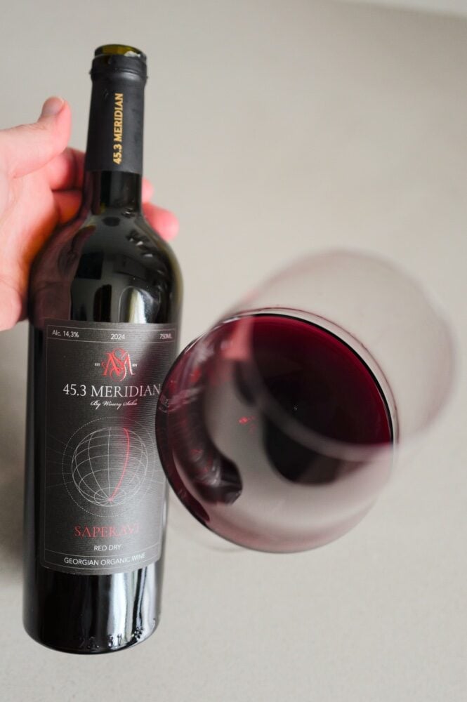 Hand holding the 45.3 Meridian Saperavi bottle beside a tilted glass of deep red wine