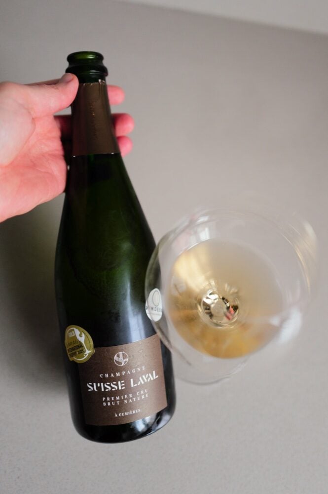 Hand holding the Suisse Laval Brut Nature bottle next to a tilted glass of champagne