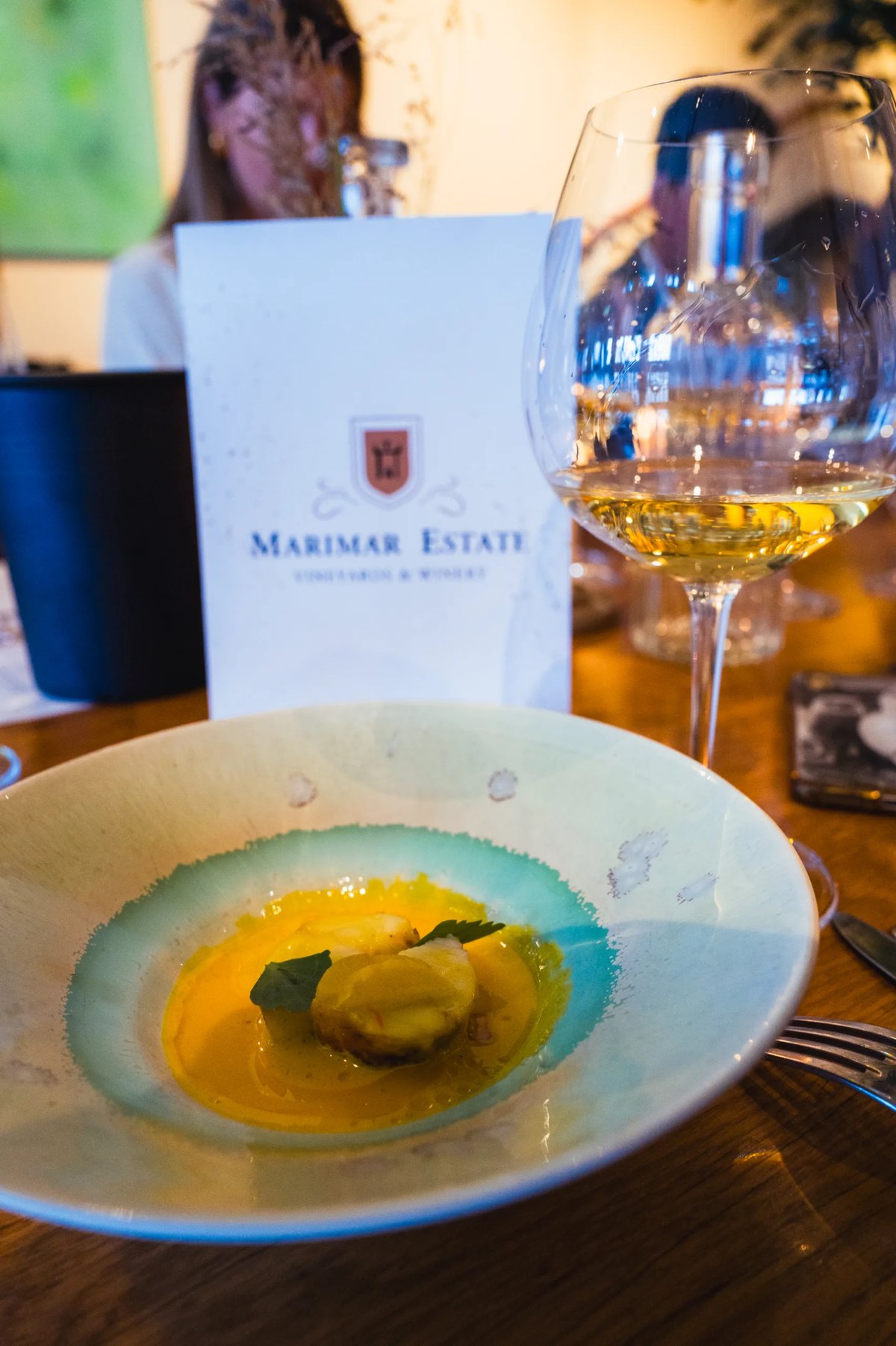 Plated course on a green-rimmed plate beside a glass of white wine and a Marimar Estate card