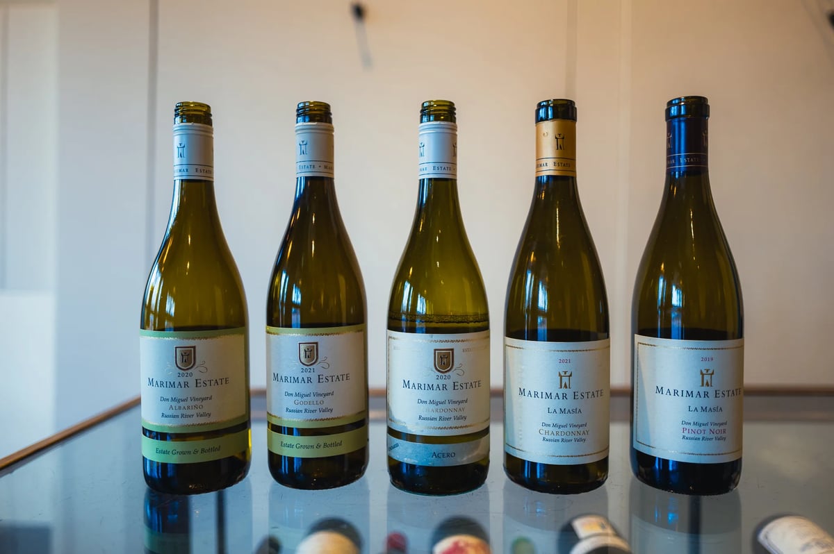 Five Marimar Estate white and red wine bottles lined up on a glass table