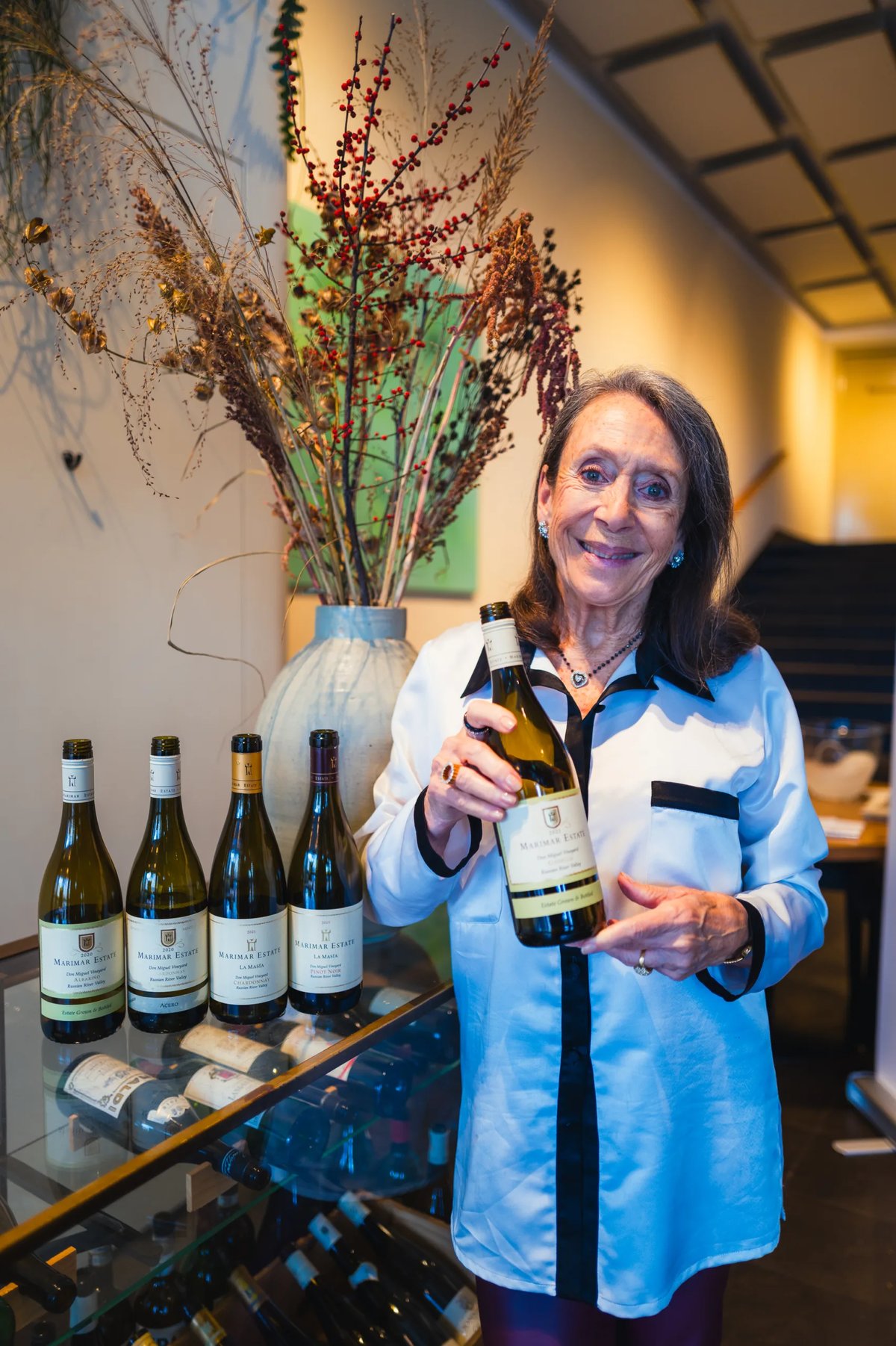 Marimar Torres presenting a Chardonnay bottle beside a lineup of her wines and dried flowers