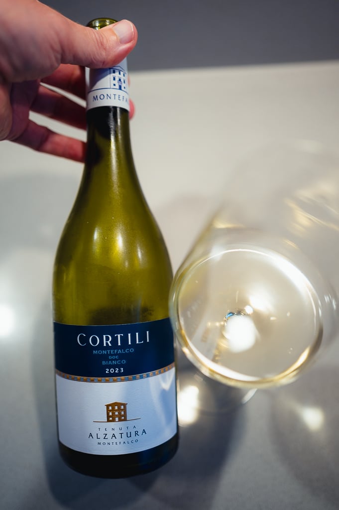 Hand holding the Cortili Montefalco Bianco 2023 bottle beside a glass of white wine seen from above