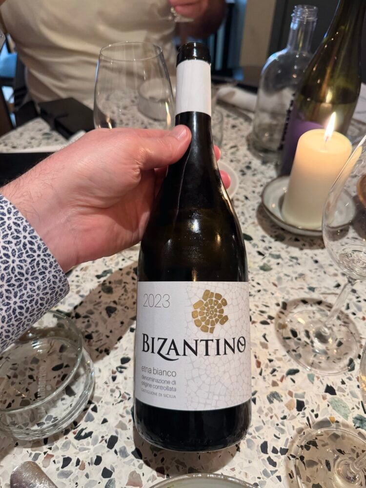 Hand holding a Bizantino 2023 Etna Bianco bottle above a candlelit terrazzo table