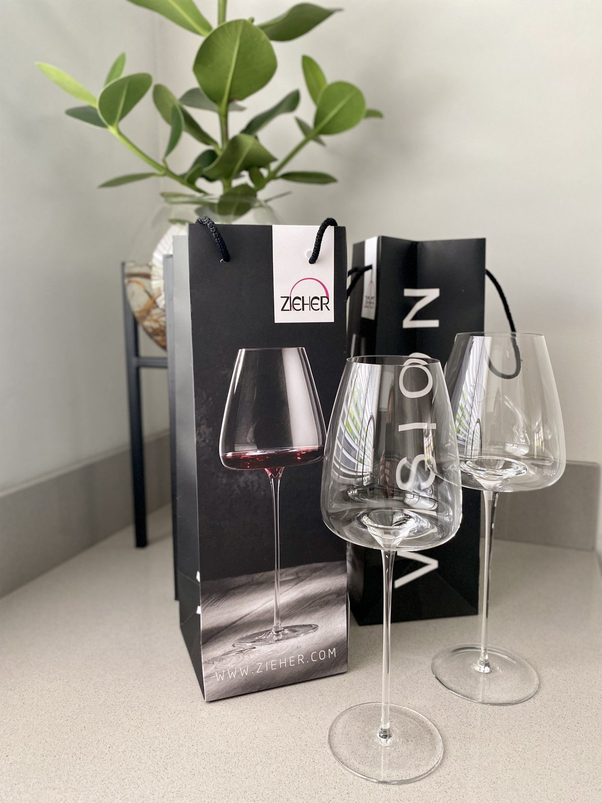 Two slender Zieher wine glasses next to black Zieher gift bags and a green plant
