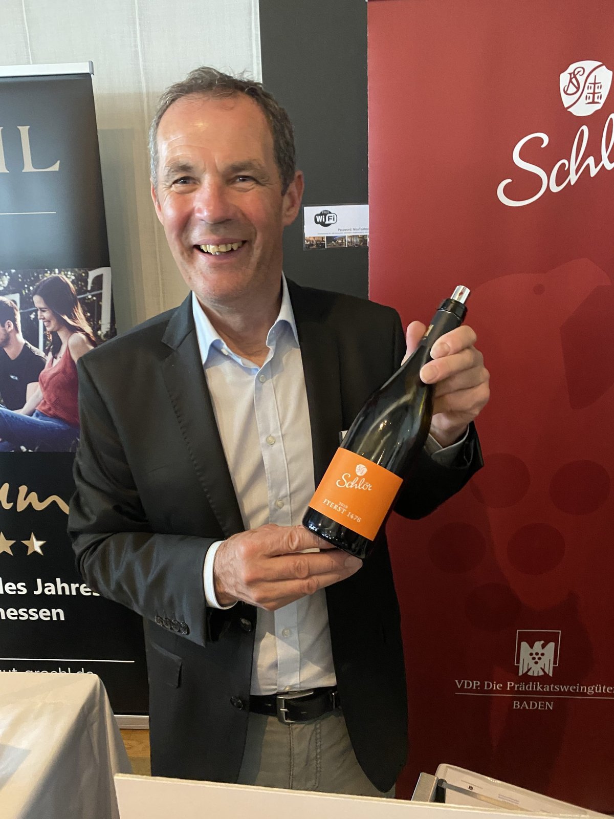 Smiling man in a blazer holding a Schlör Fyerst bottle beside a red Schlör VDP Baden banner
