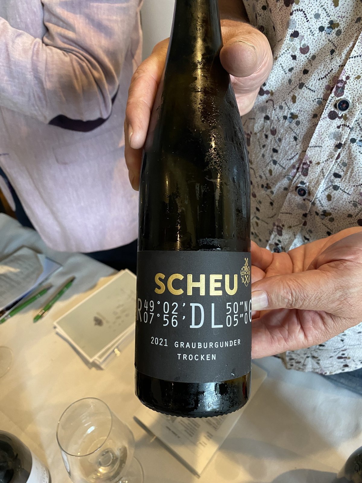 Two people holding a black Scheu DL 2021 Grauburgunder Trocken bottle at the tasting table