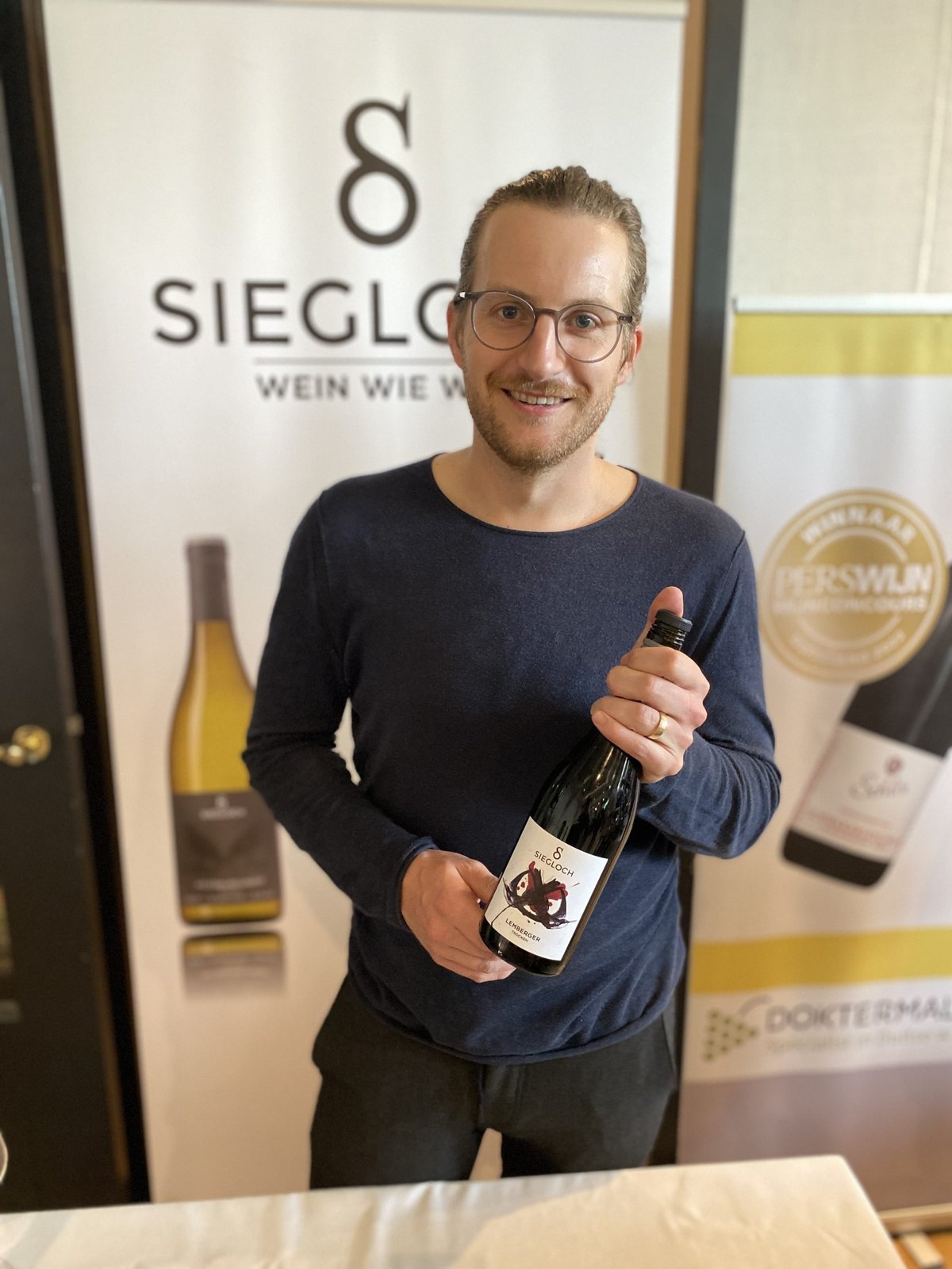 Smiling man holding a Siegloch Lemberger bottle in front of a Siegloch winery banner