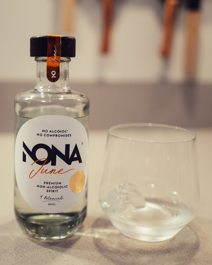 Clear NONA June non-alcoholic spirit bottle beside a tumbler with a clear drink over ice