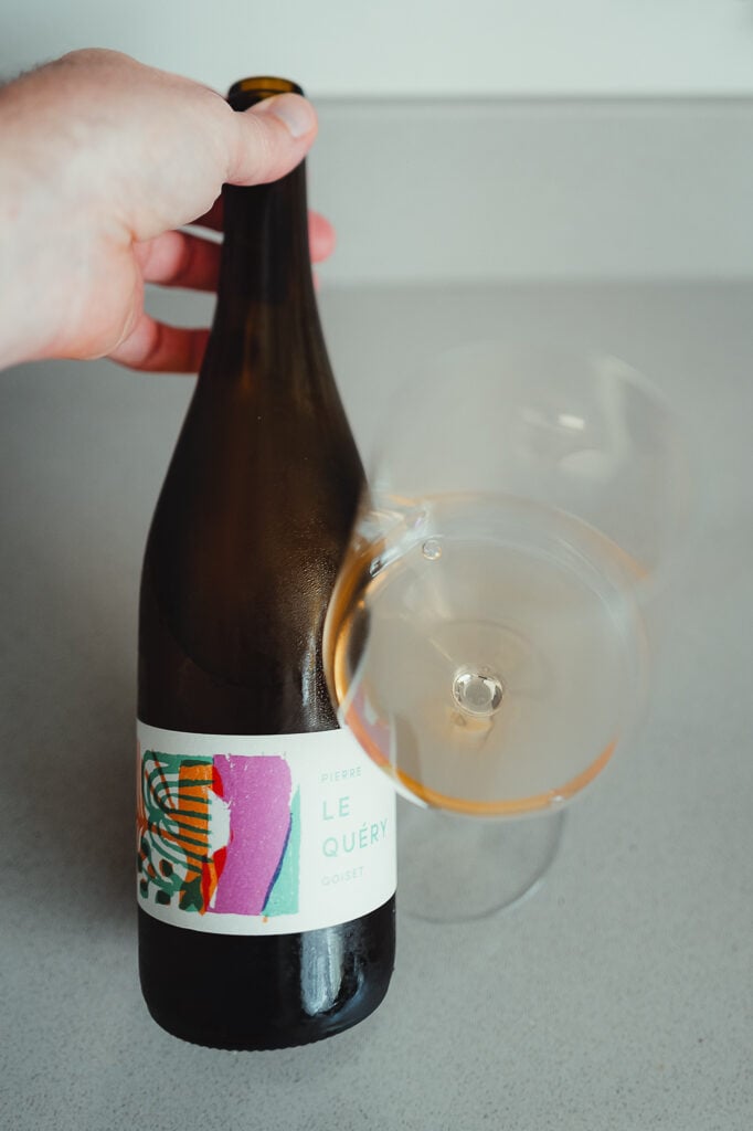 Hand holding the Pierre Goiset Le Quéry bottle next to a tilted glass of orange wine