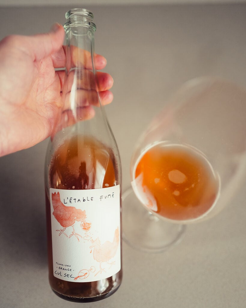 Hand holding the Cul Sec L'étable Fumé bottle with a hen-illustrated label beside a glass of orange drink