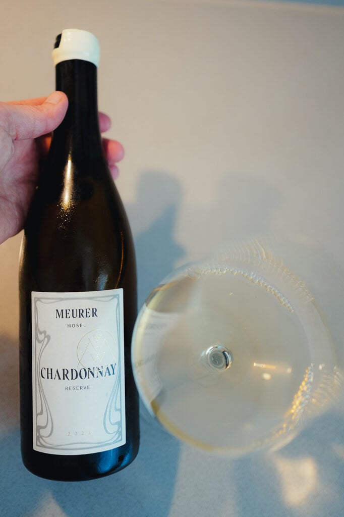 Hand holding the Meurer Mosel Chardonnay Reserve bottle beside a glass of white wine seen from above