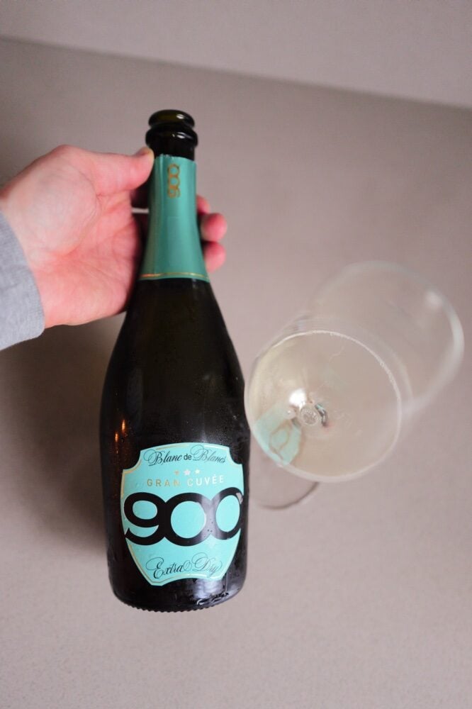 Hand holding the teal 900 Gran Cuvée Blanc de Blancs bottle next to a glass