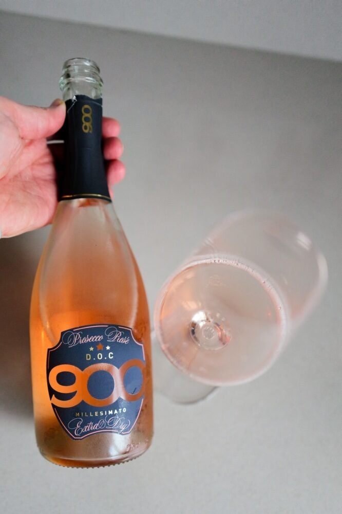 Hand holding the 900 Prosecco Rosé DOC bottle next to a glass of pink sparkling wine