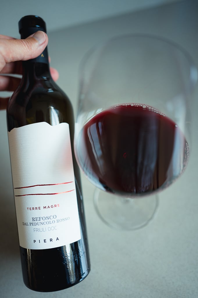 Hand holding the Piera Refosco dal Peduncolo Rosso bottle next to a glass of red wine