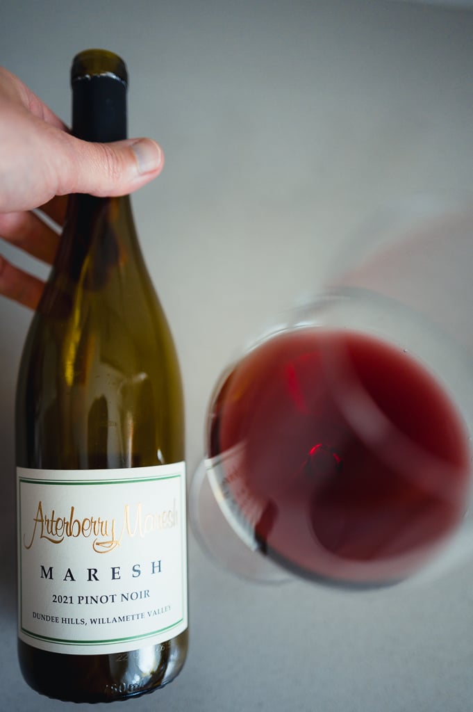 Hand houdt fles Arterberry Maresh 2021 Pinot Noir naast een glas rode wijn van bovenaf