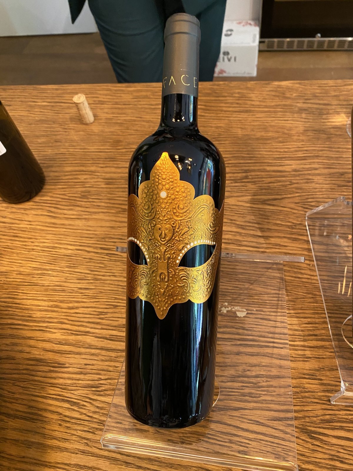 Dark red wine bottle with an ornate gold mask label resting on a wooden table