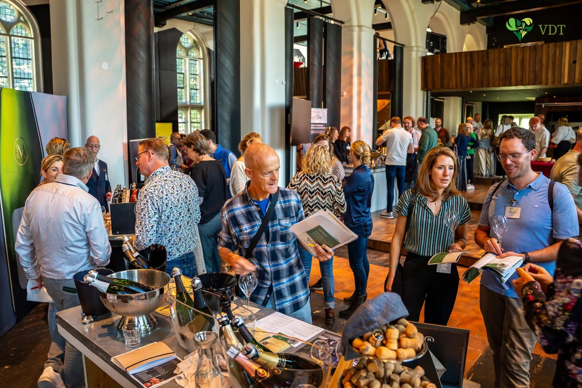 Busy champagne tasting event in a former church hall, with attendees browsing tables of bottles and ice buckets under the VDT banner